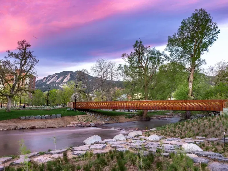 Bridge over a river in a park, with Flatirons mountains in the background under a colorful sunset.