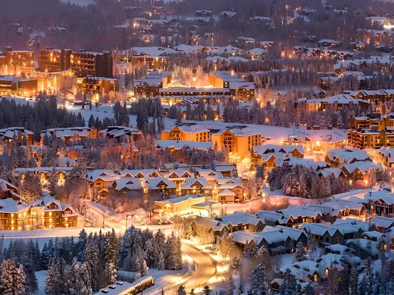 Snow-covered mountain town at night, buildings with warm lights, trees, and a twilight sky.