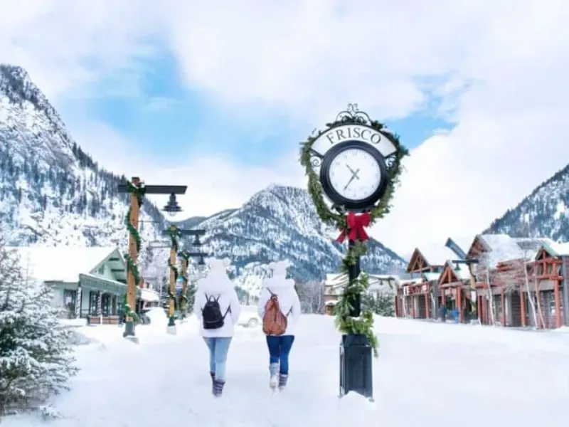 Two people walk toward a clock in Frisco, CO, in the snow, past shops and mountains.