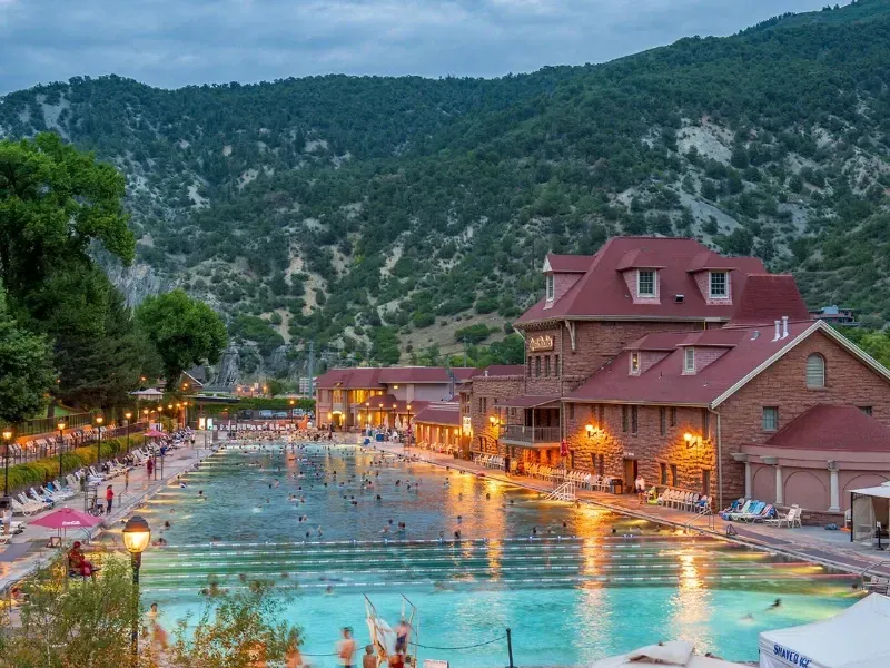 People swim in a large outdoor pool surrounded by brick buildings at a hot springs resort, with mountains in the background.