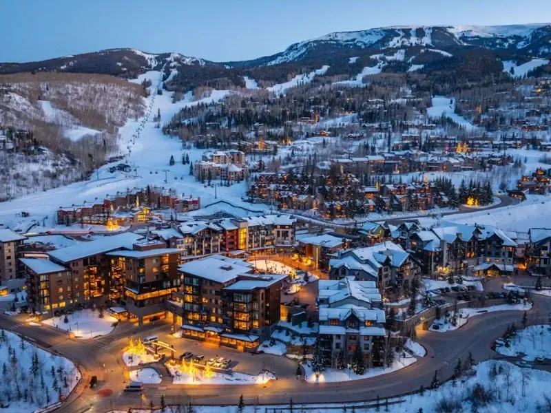 Snowy ski resort town at dusk, illuminated buildings nestled at base of mountains.