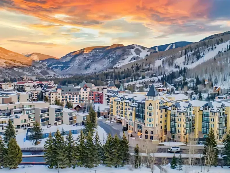 Snowy mountain town with colorful sky at sunset. Buildings and ski slopes are visible.