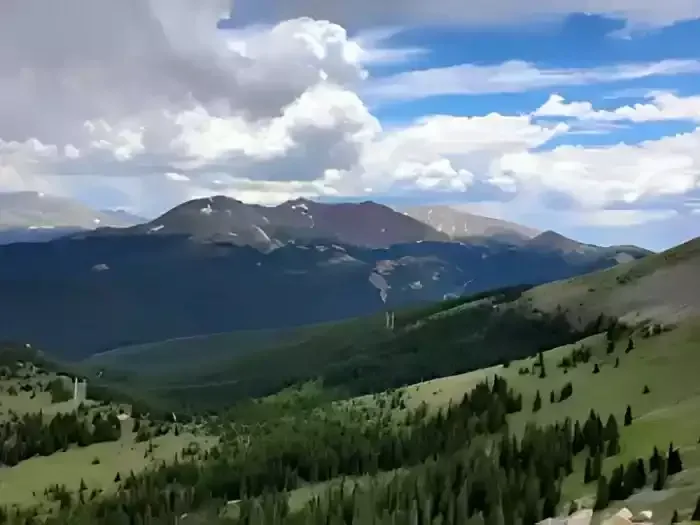 Mountain valley with green slopes, dark forest, and snowy peaks under a cloudy blue sky