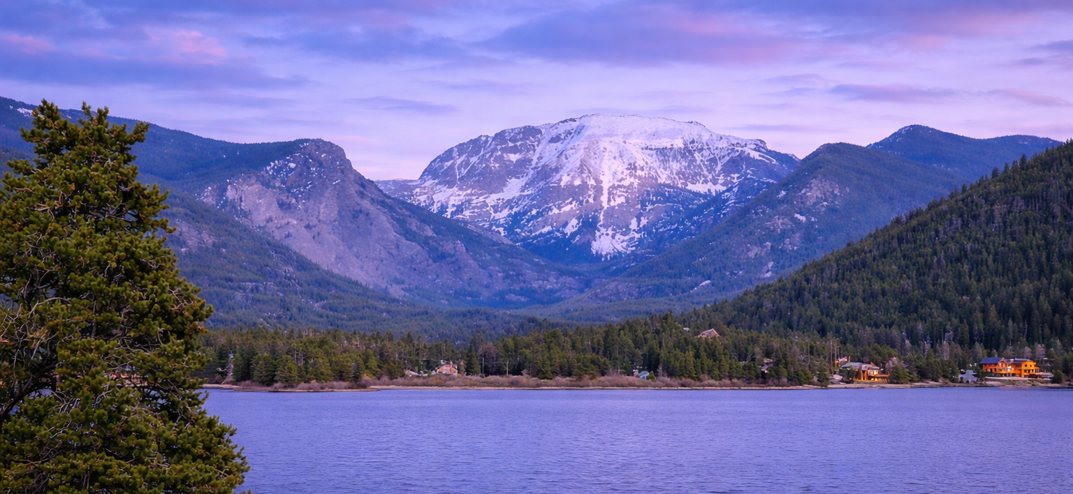 Snow-capped mountains tower over a deep blue lake surrounded by a dense evergreen forest under a soft twilight sky.