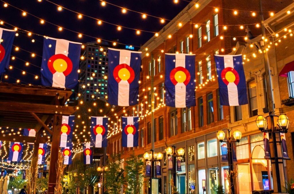 Colorado state flags hang vertically above a Denver, CO street, illuminated by festive string lights at night. Colorado state flags hang vertically above a Denver, CO street, illuminated by festive string lights at night.