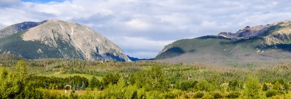 Wide mountain valley with green forest, rocky peaks, and a partly cloudy sky