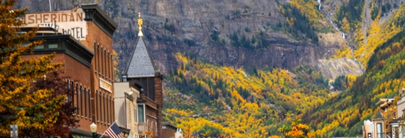 Mountain town street with a hotel sign, church steeple, and autumn trees against rocky cliffs