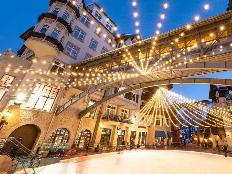 Buildings with a bridge over an ice rink, strung with holiday lights. Evening scene.