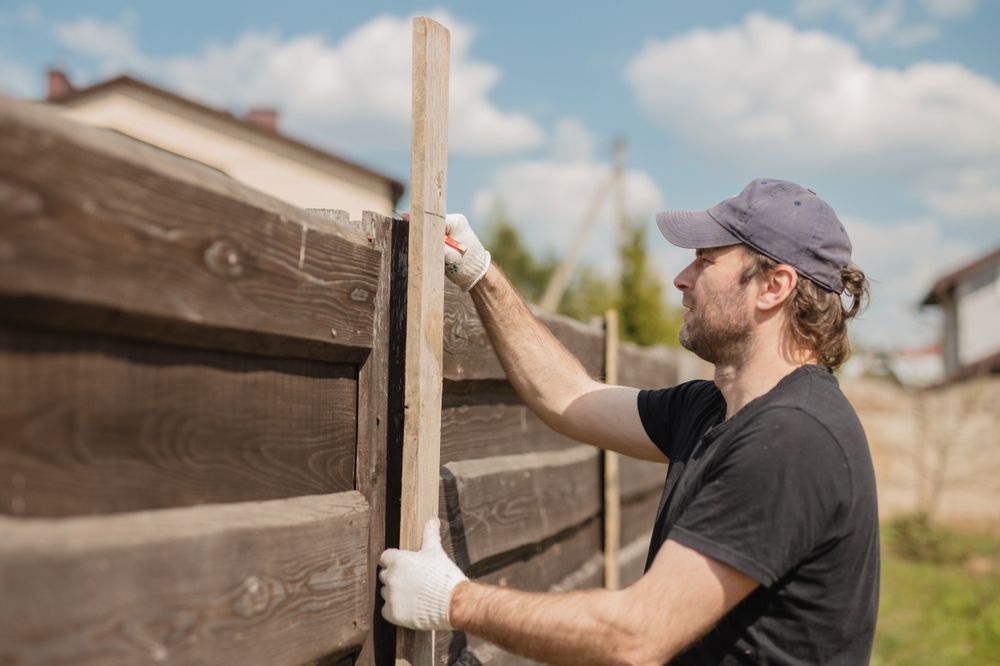 Man in a cap and gloves adjusting a wooden post next to a brown wooden fence.