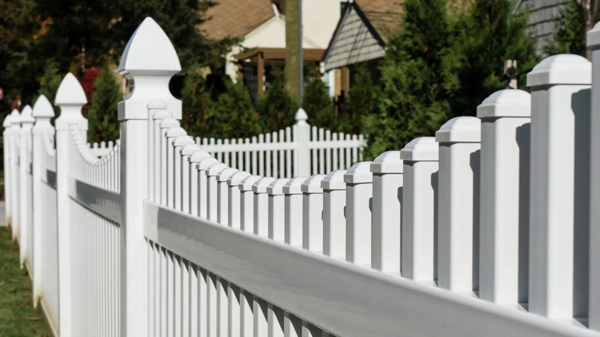 White picket fence in front of green grass and trees with houses in the background.