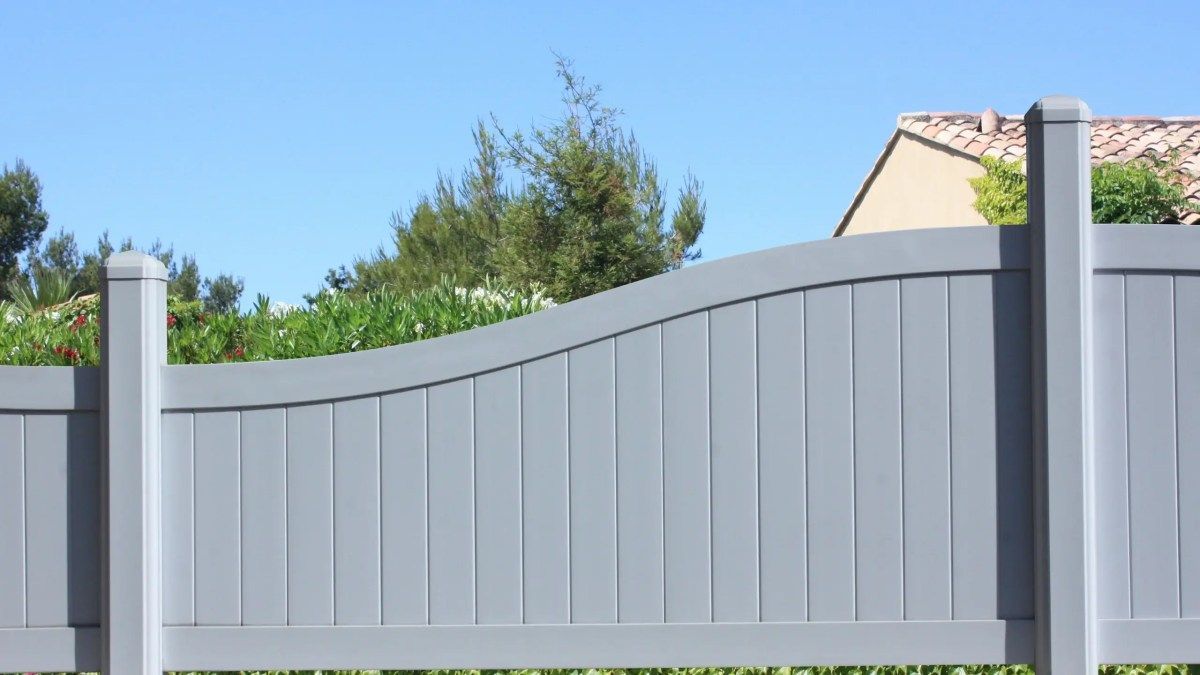 Gray wooden fence with curved top, against blue sky and greenery.