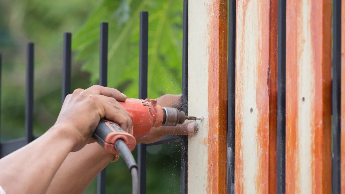 Person using a power drill to attach a wooden panel to a black metal fence.