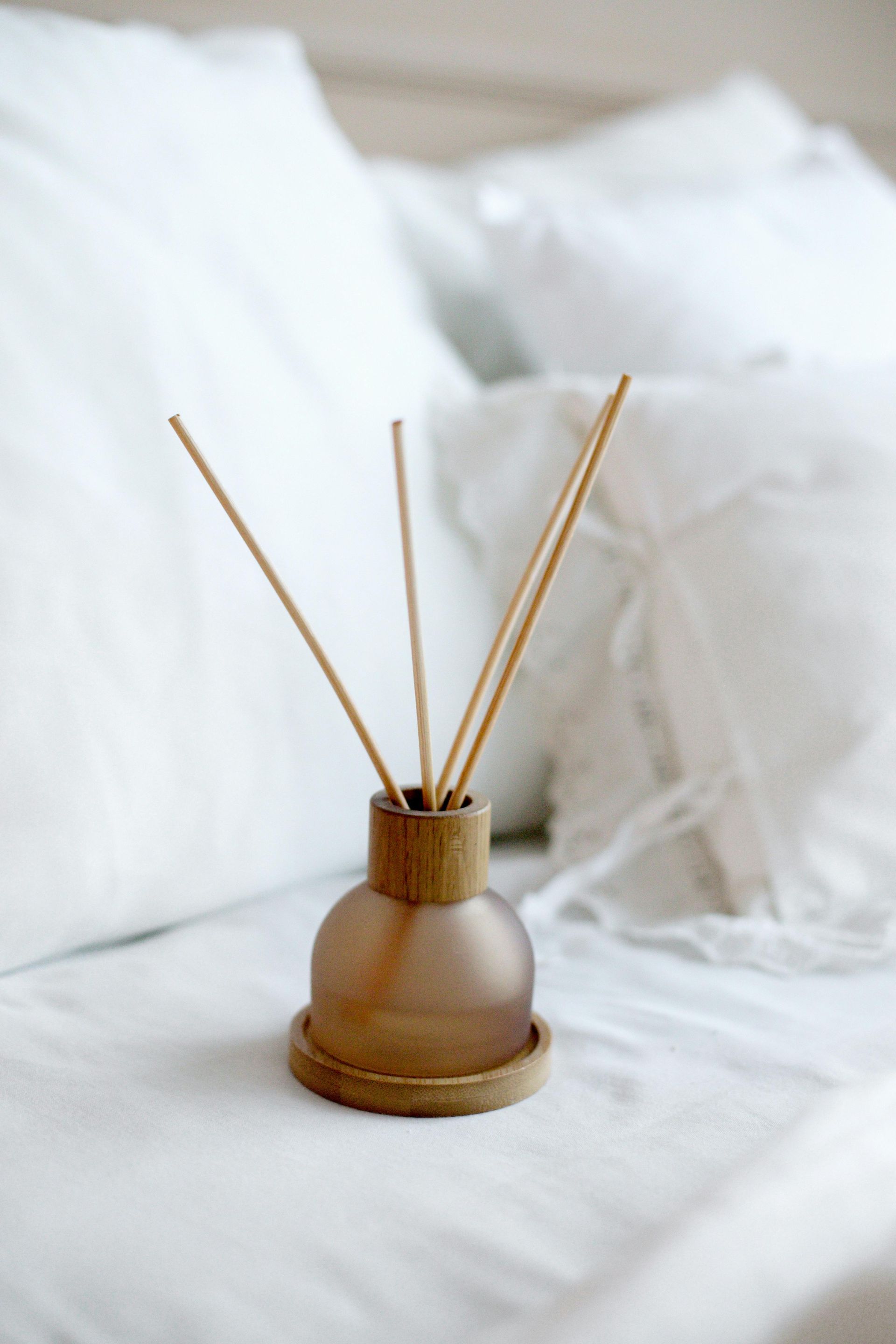 Diffusore a bastoncini marrone con bastoncini su un letto bianco tra cuscini bianchi.
