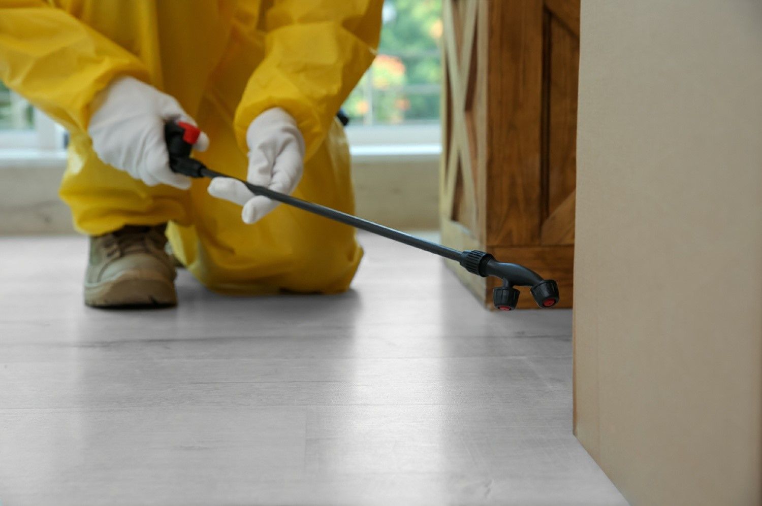 Worker in yellow protective suit spraying pesticide along a floor edge with a wand sprayer