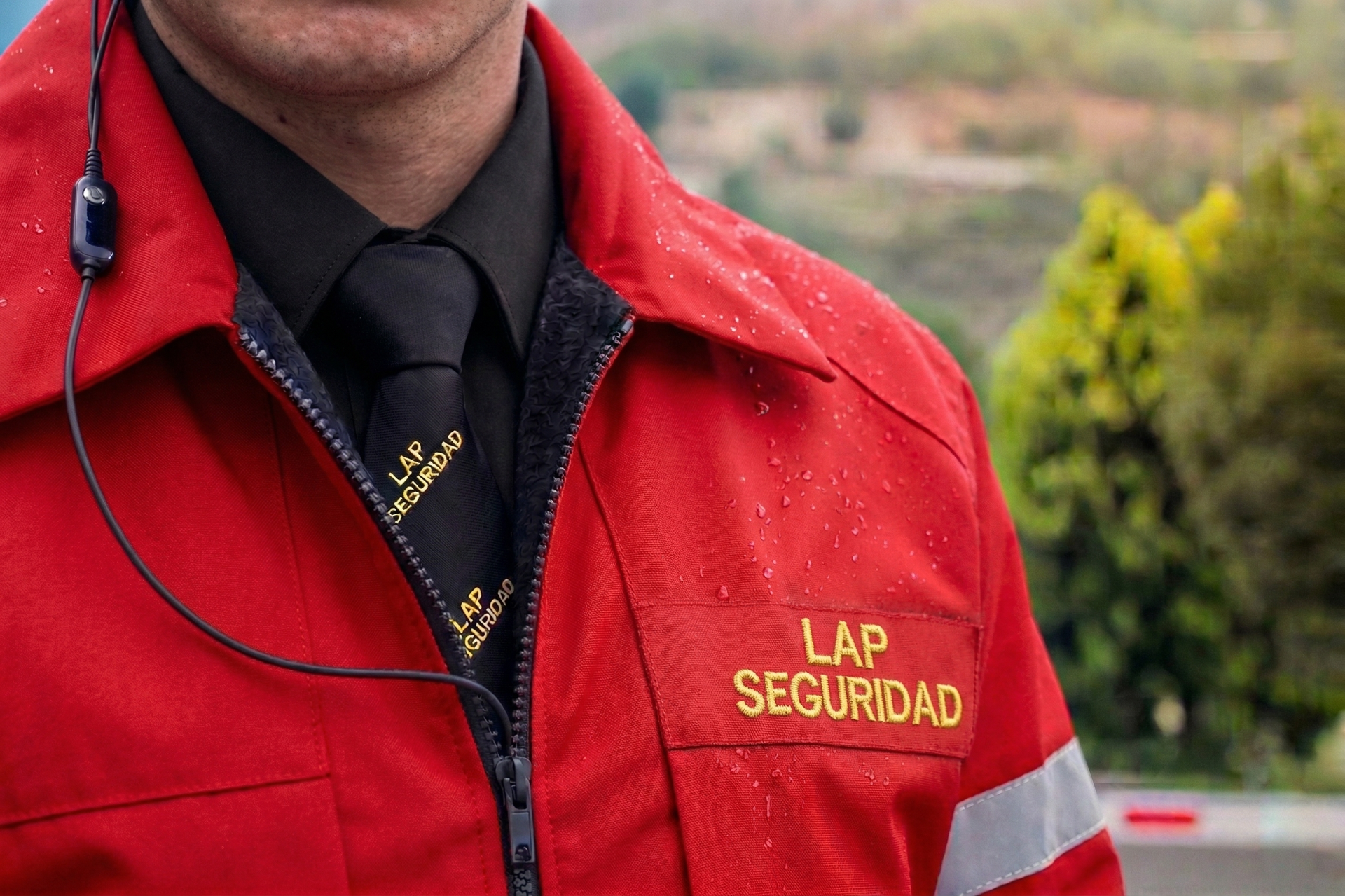Persona con chaqueta roja de seguridad con el texto “LAP SEGURIDAD” y corbata negra, al aire libre.