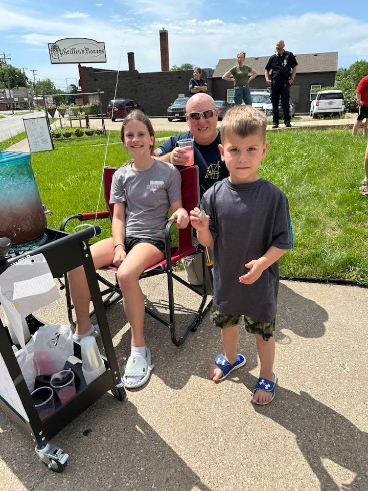 A man and two children sit near a drink cart on a sunny day. The children are smiling, and the setting appears to be outdoors.