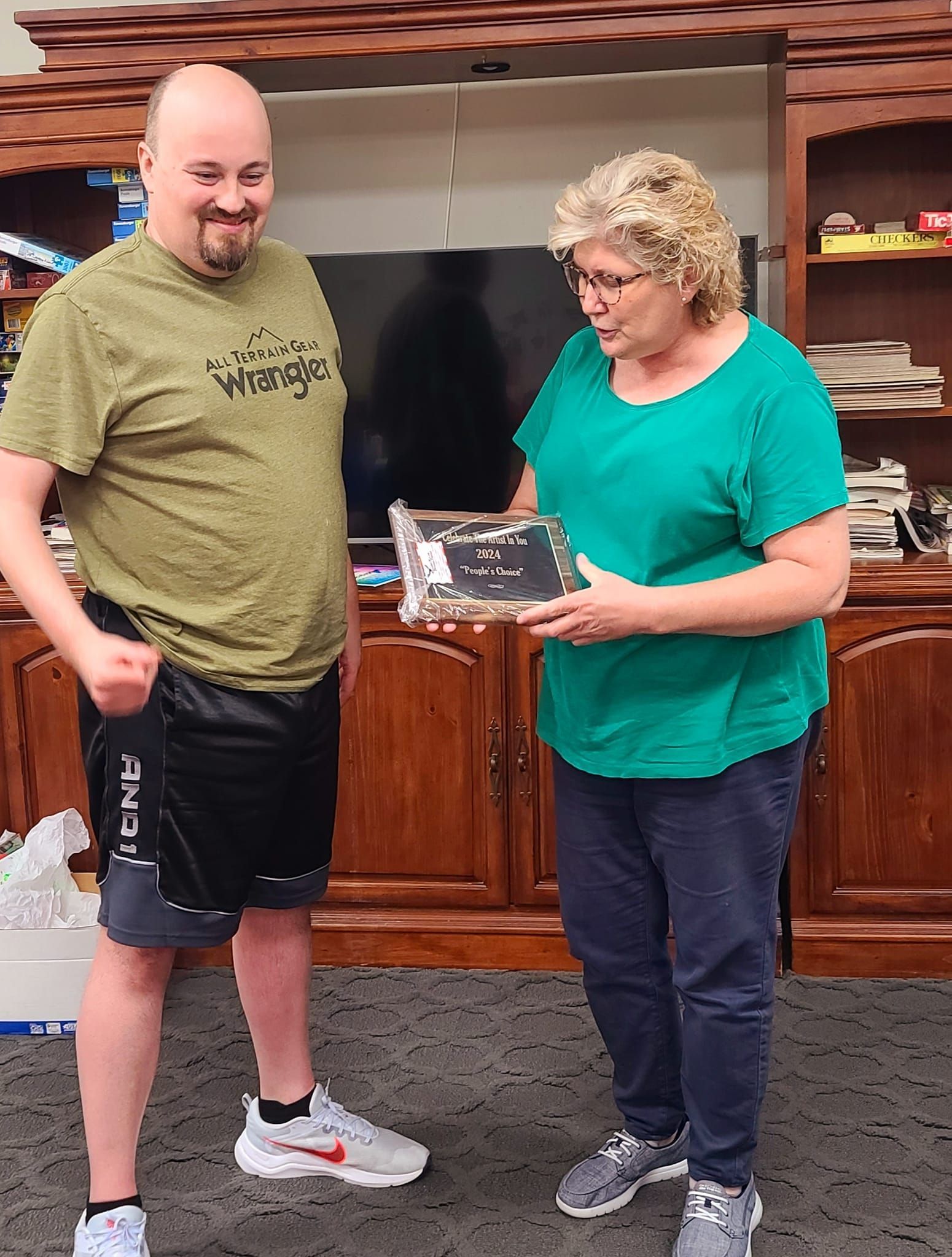 A woman hands a framed item to a man in a home setting. The man wears a green shirt and shorts, the woman a teal shirt and jeans.