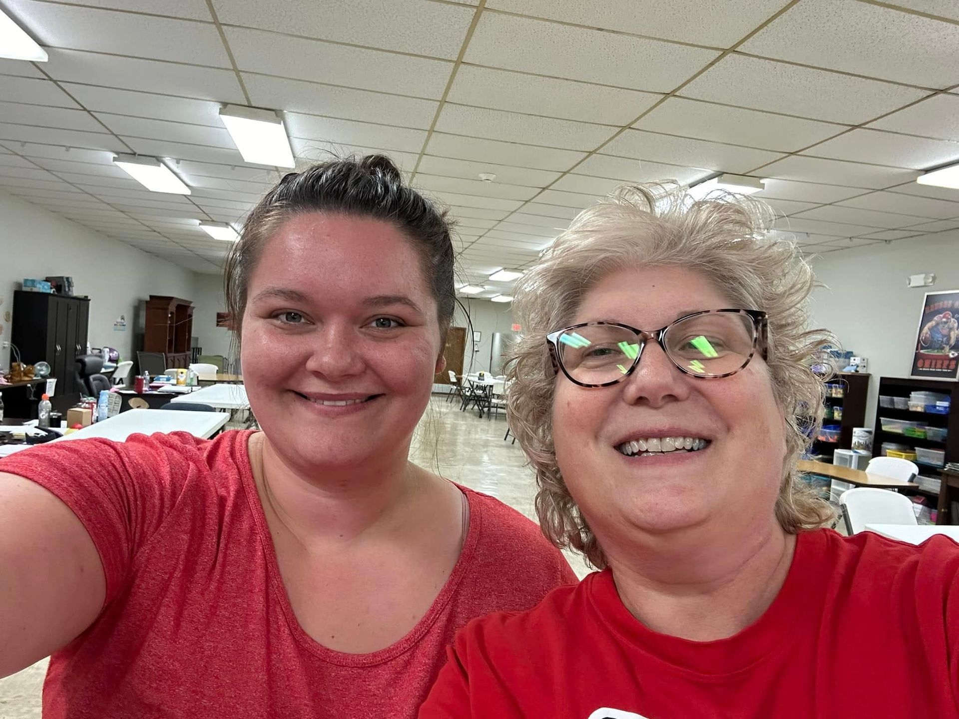 Two women smiling close to camera in a brightly lit room. One in red shirt, glasses, and short hair; other in a red shirt with dark hair.