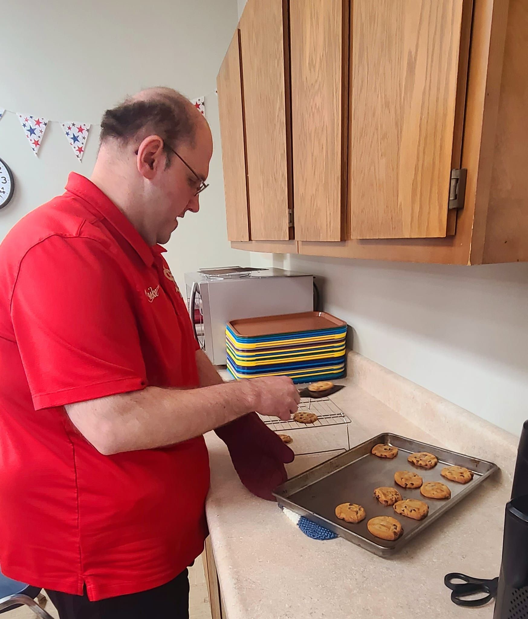 A man wearing a red shirt and glasses takes cookies from a baking sheet in a kitchen.