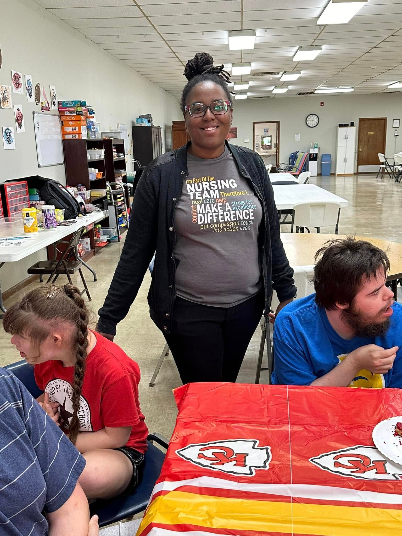 A woman wearing glasses and a black cardigan stands with a group of people, including a girl in a red shirt and a man with a beard.