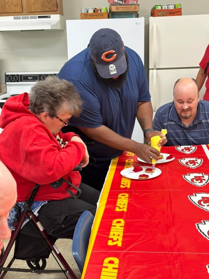A man in a Bears hat puts mustard on a plate for a woman and another man at a table covered in a Chiefs tablecloth.