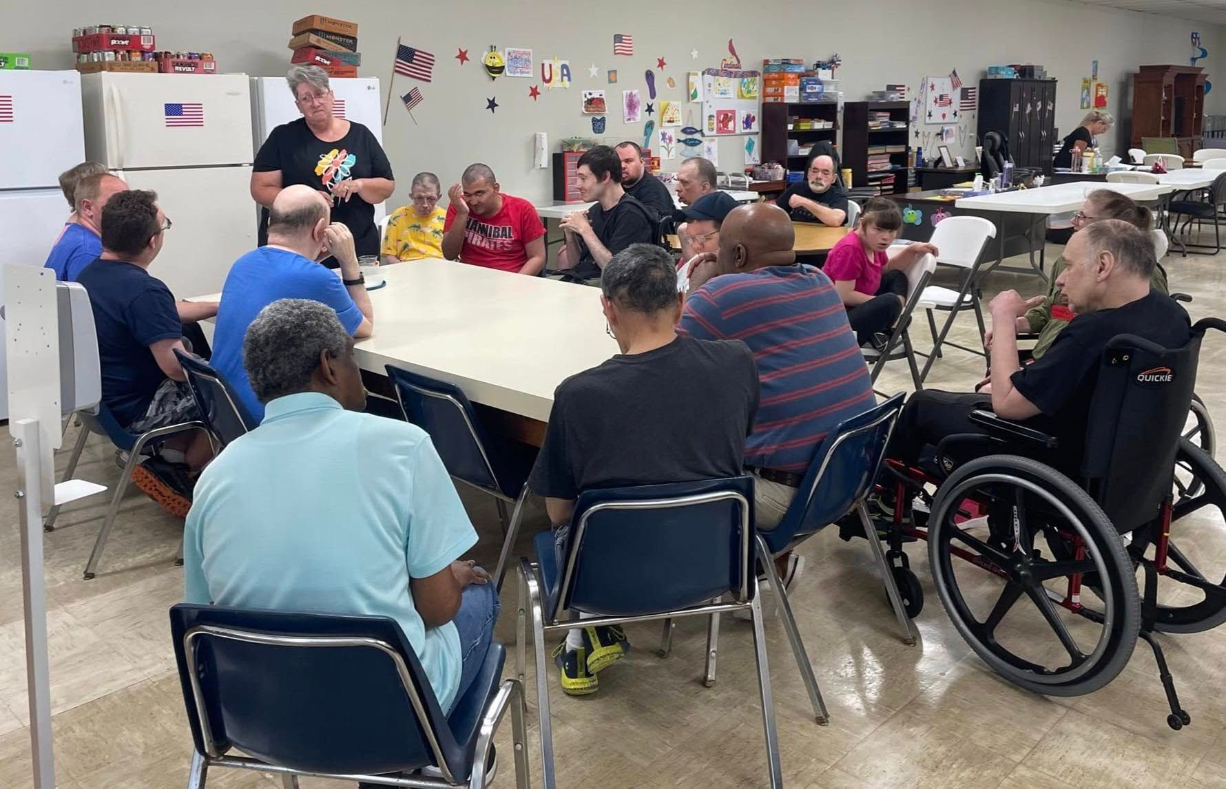 Group of people in a room seated around a table, with a woman standing and addressing them. Some use wheelchairs.