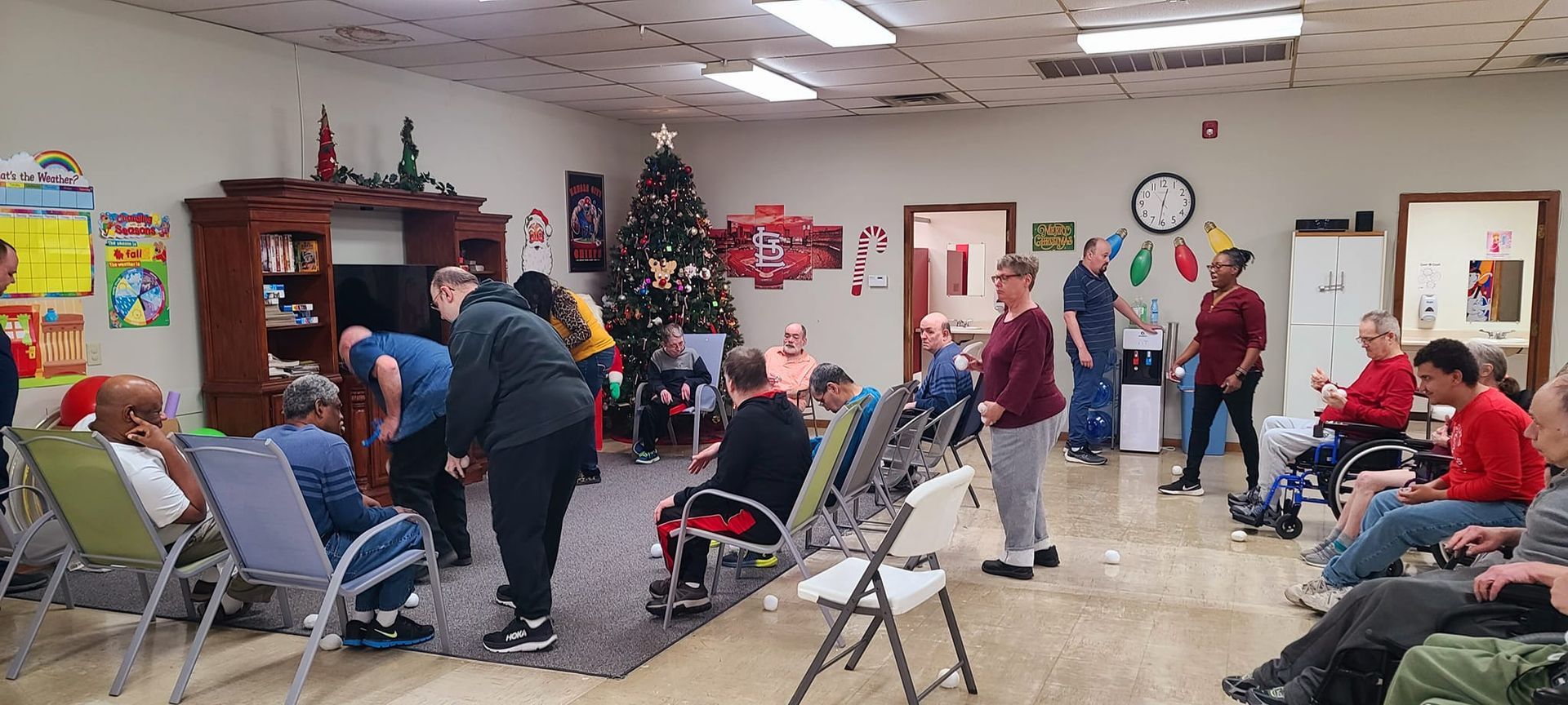 A group of people in a decorated room, possibly a community center. Some are in wheelchairs or seated, others are standing and some are participating in activities. A Christmas tree adds to the festive atmosphere.