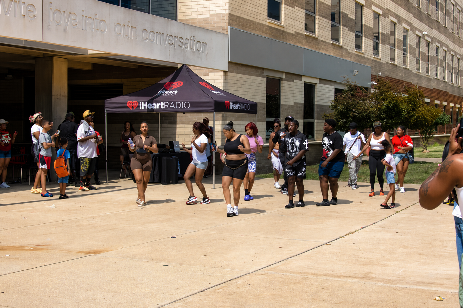 People standing in line outside a building near a black tent on a sunny day