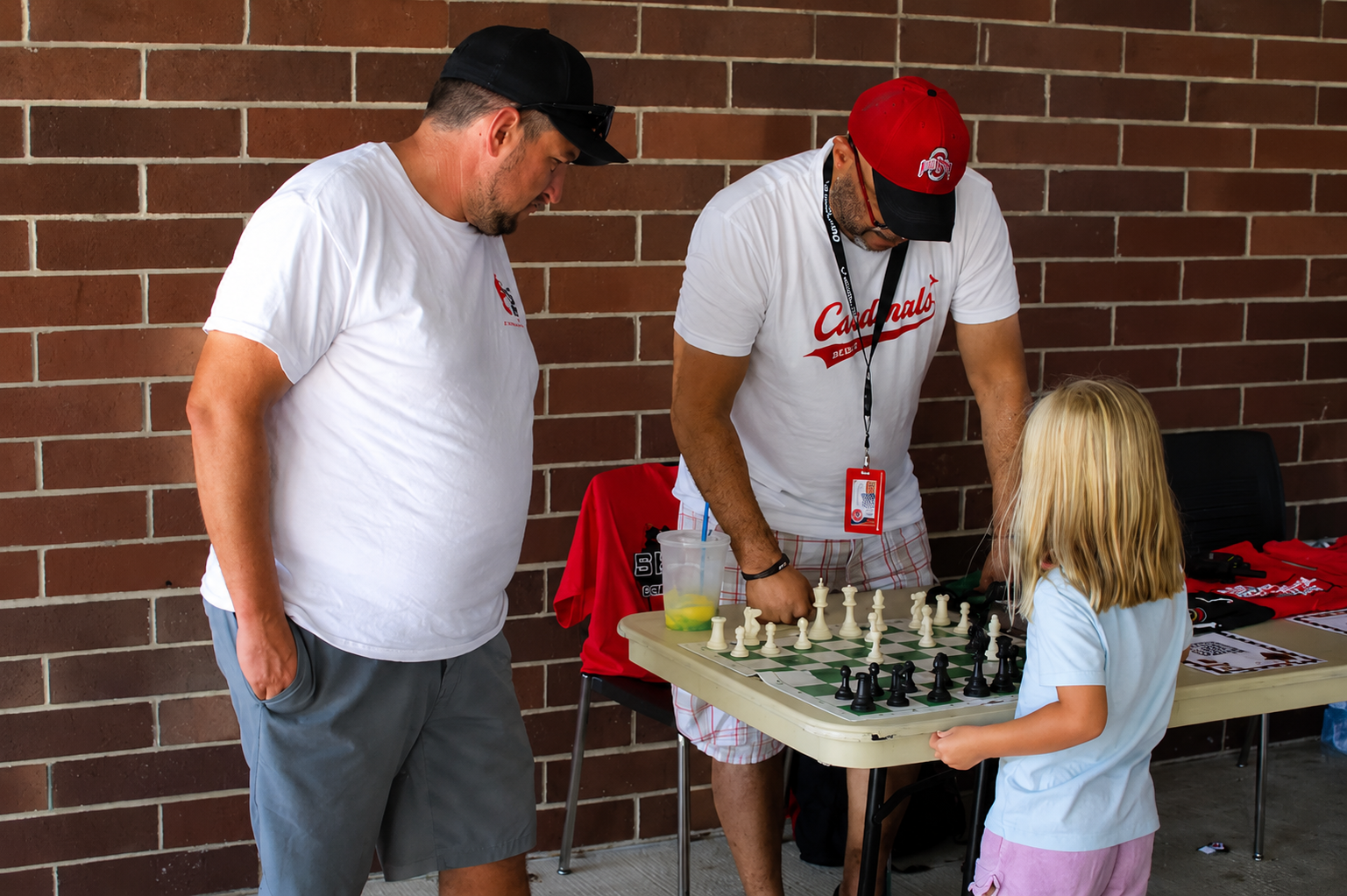 Three people at an outdoor table, playing chess beside a brick wall.