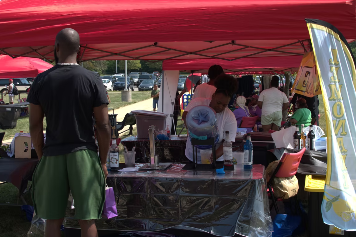 Two people stand behind a table with food items. Others stand nearby. Cloudy, outdoor setting.