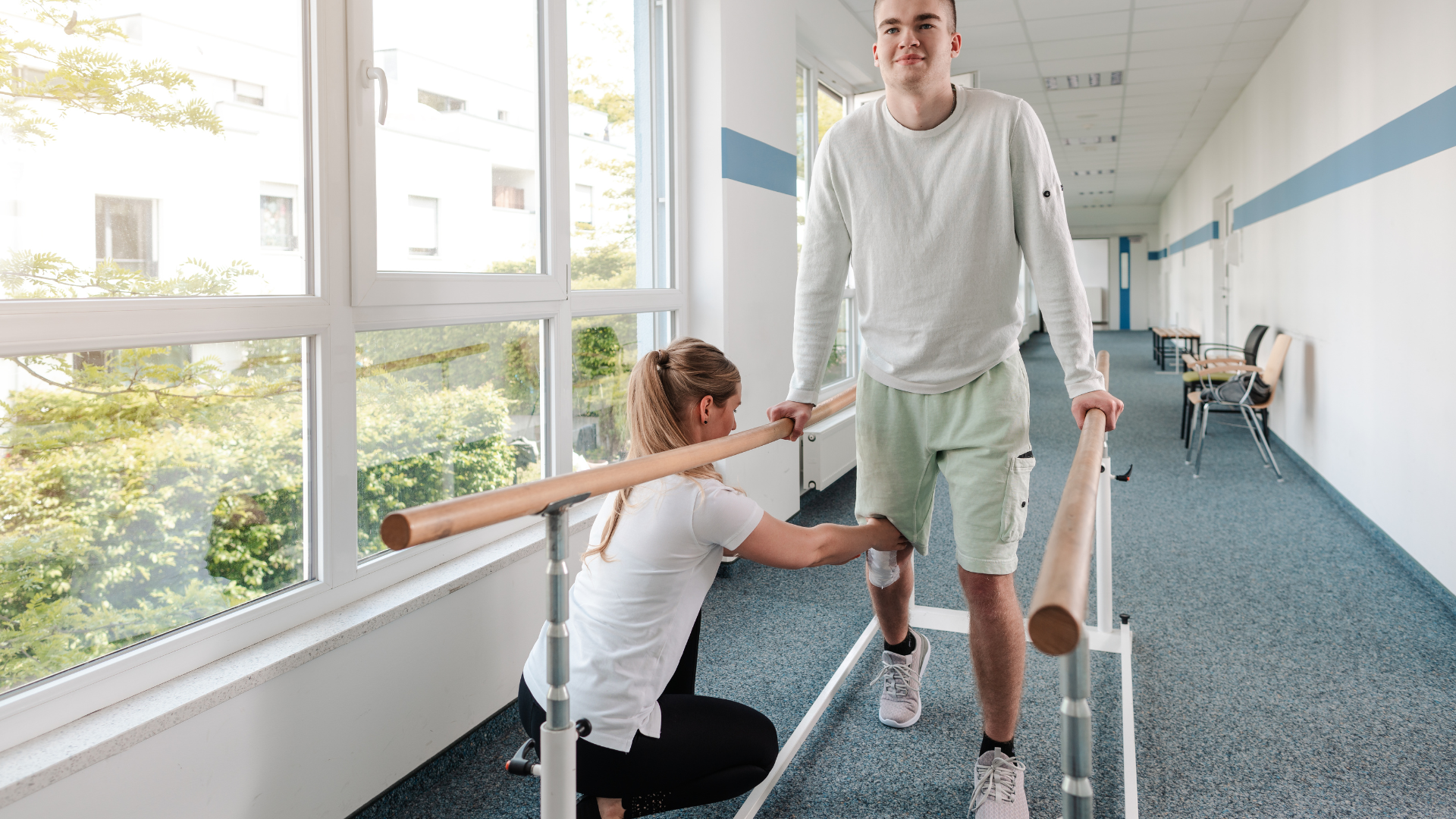 A woman is helping a man walk on parallel bars in a hallway.