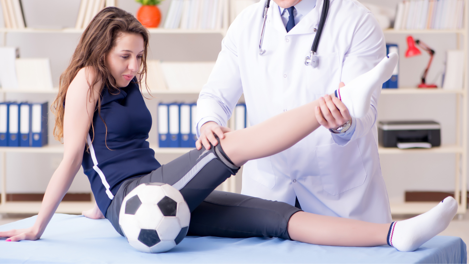 A woman is sitting on a bed while a doctor examines her leg with a soccer ball.