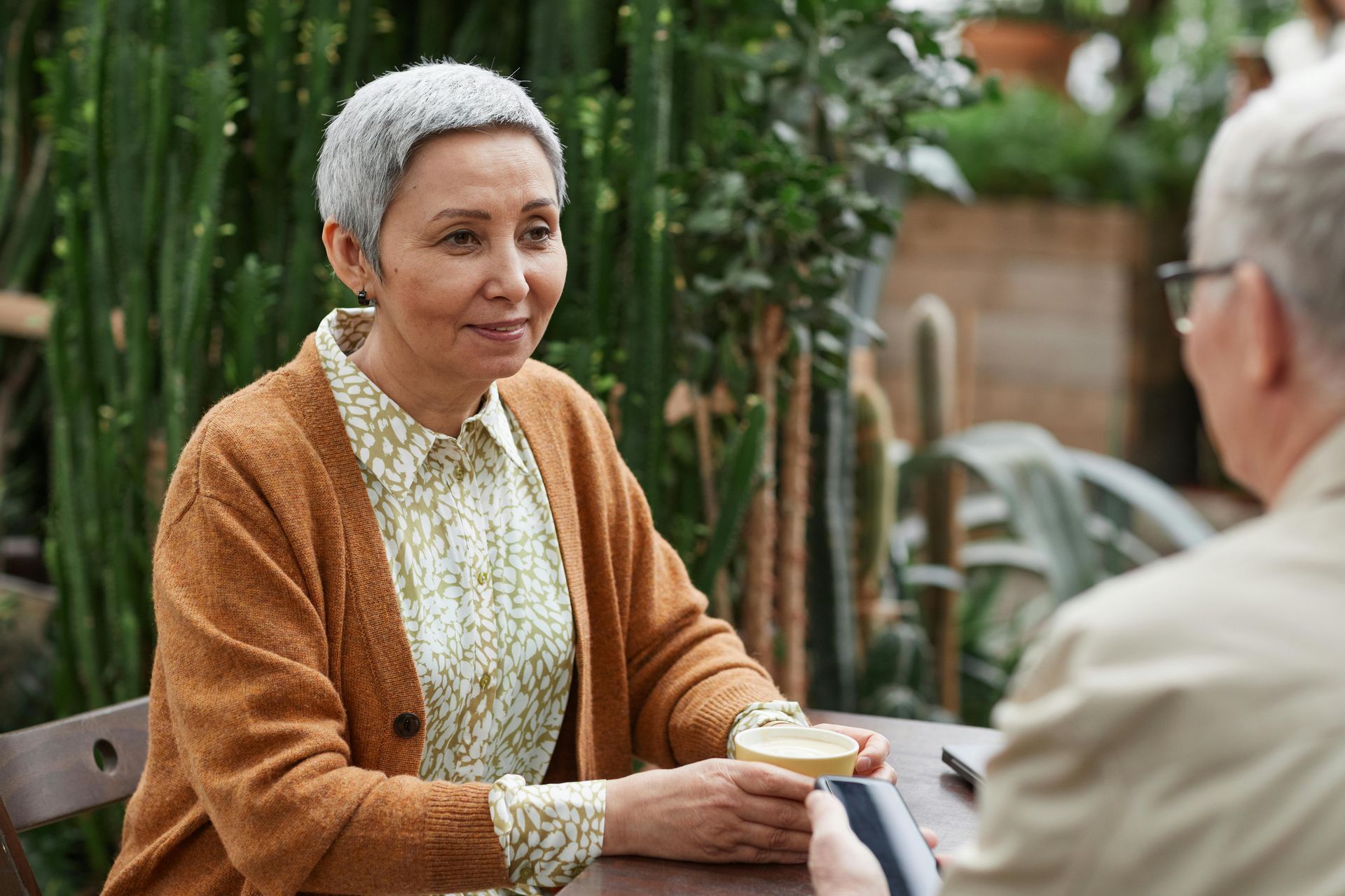 Woman with short gray hair smiles, seated at a table outside, holding a drink and looking at another person.