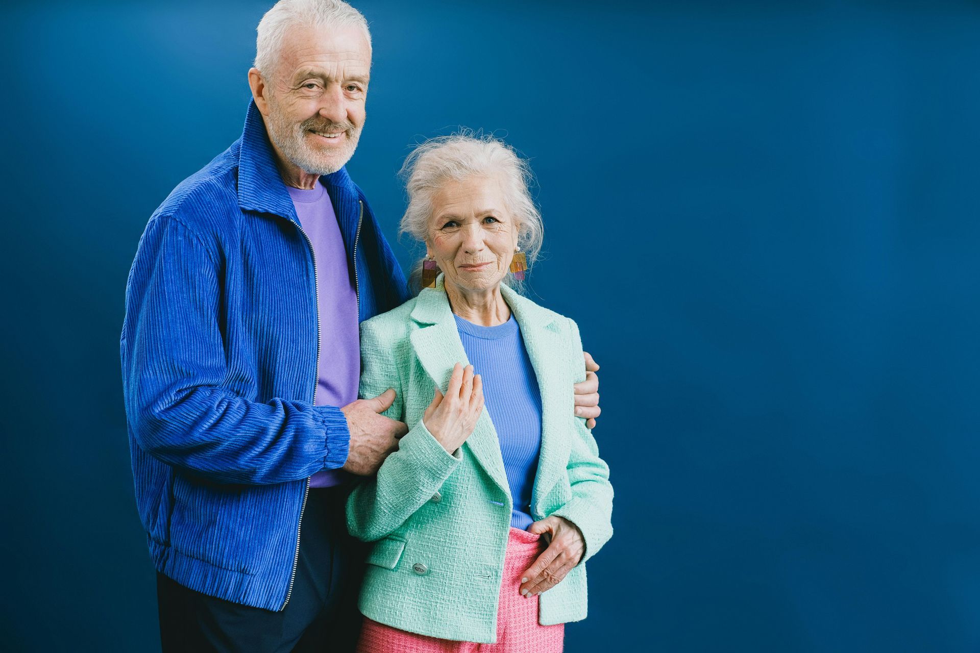 Older couple posing, man in blue jacket, woman in green jacket, smiling, blue background.
