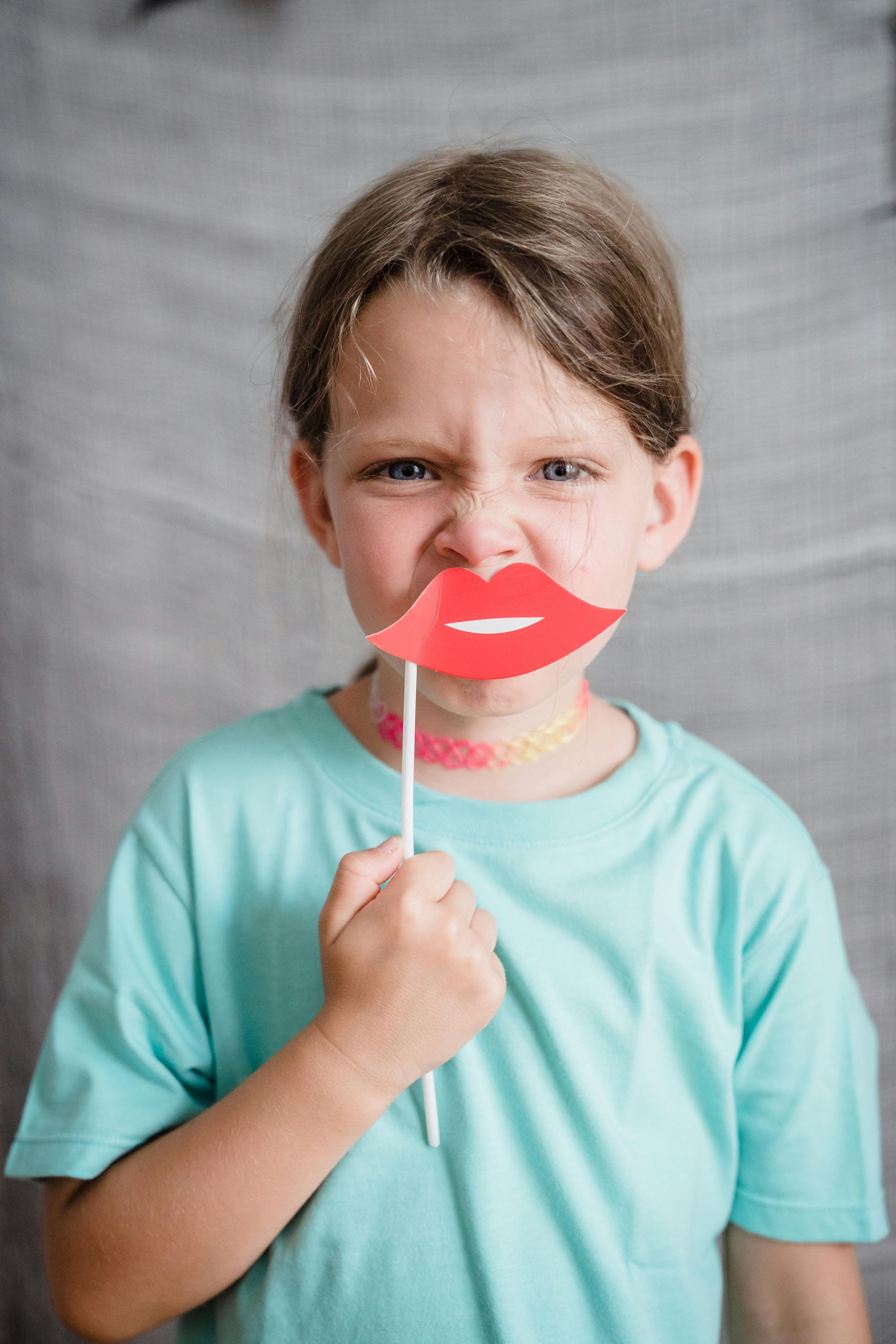 Girl with light blue shirt holding a red lip prop, making a face. Gray backdrop.