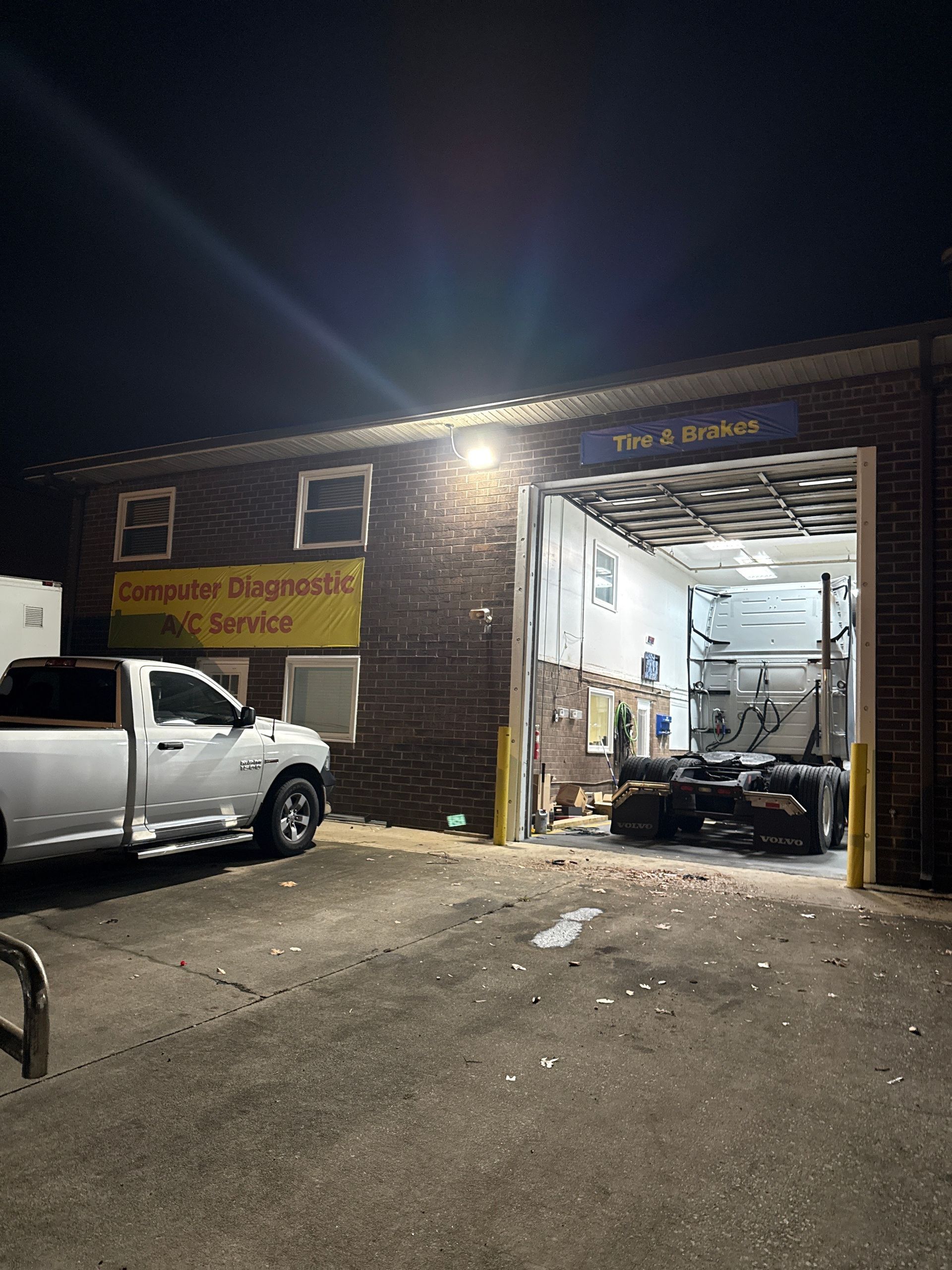 Nighttime view of an auto repair garage with a white pickup truck parked outside and tools visible inside the open bay.