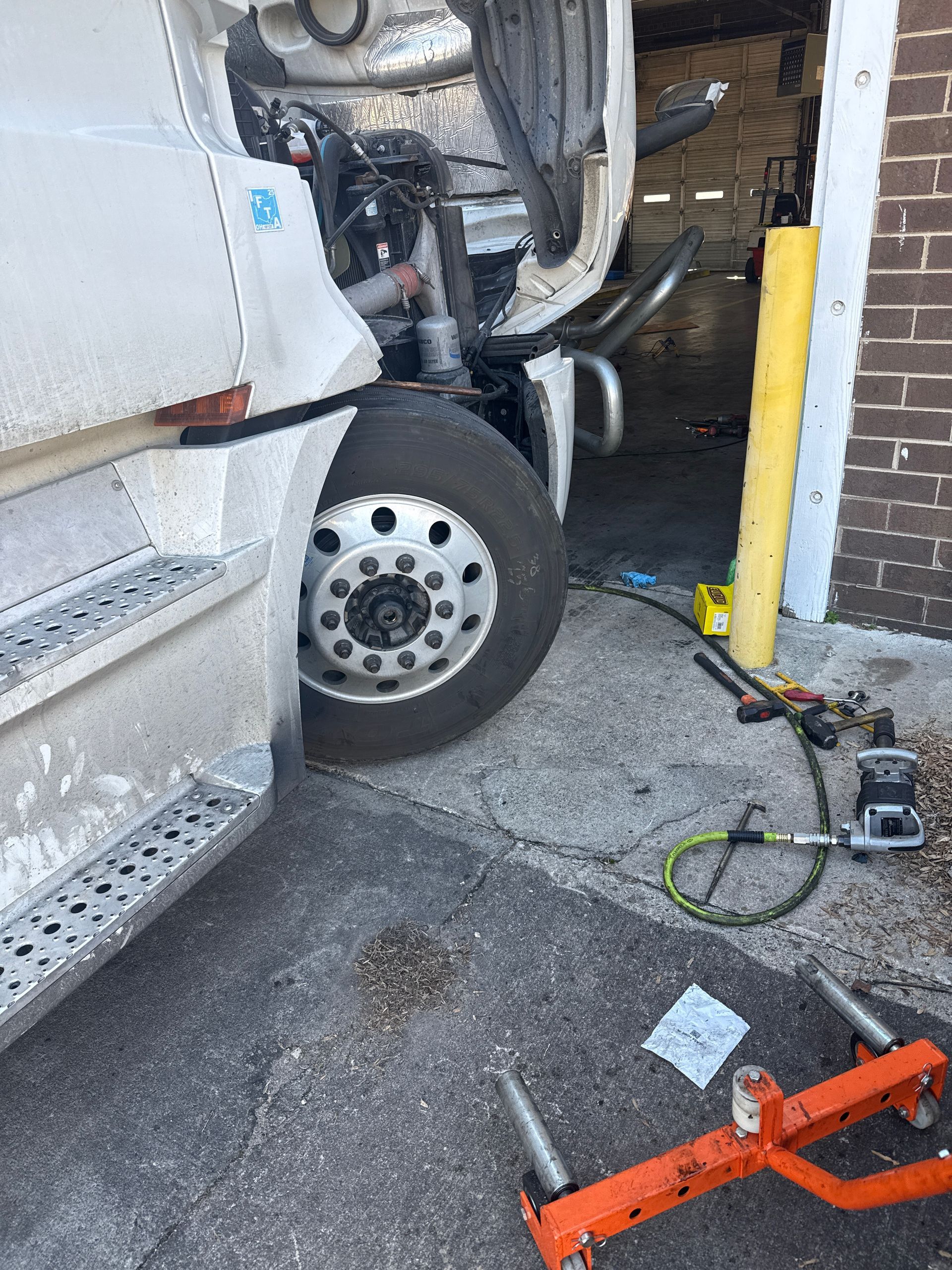 Truck wheel and exposed engine beside a curb, with a yellow bollard and tool on the pavement.