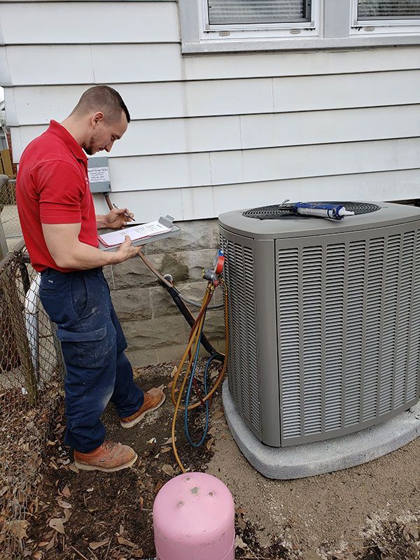 A technician in a red shirt and blue pants records readings from an AC unit connected to a pink refrigerant tank.