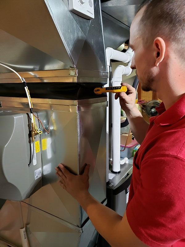A person in a red shirt uses a digital multimeter to inspect a metal HVAC unit in a utility room.