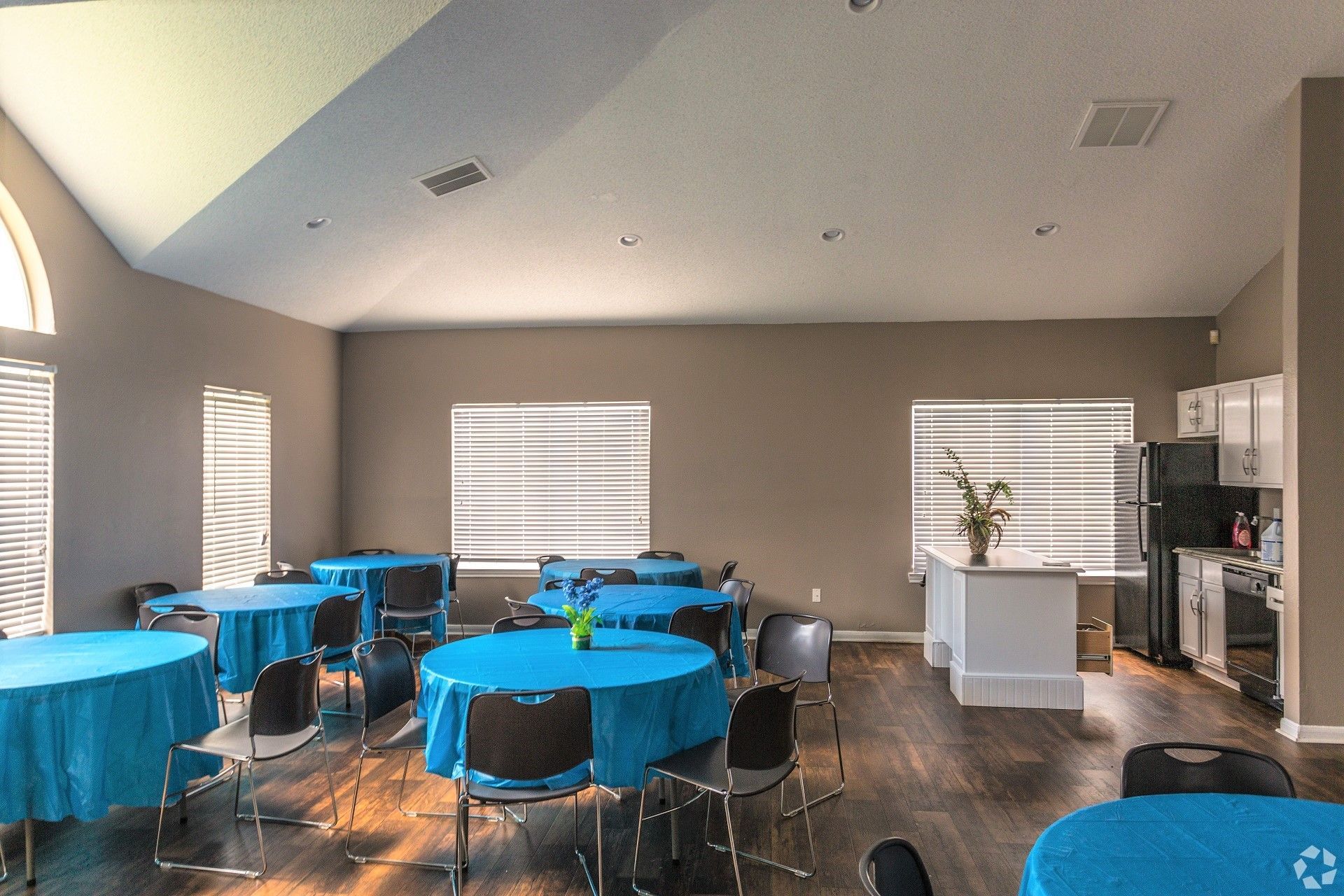 A brightly lit room with round tables covered in blue tablecloths, chairs, and a kitchen area in the background.