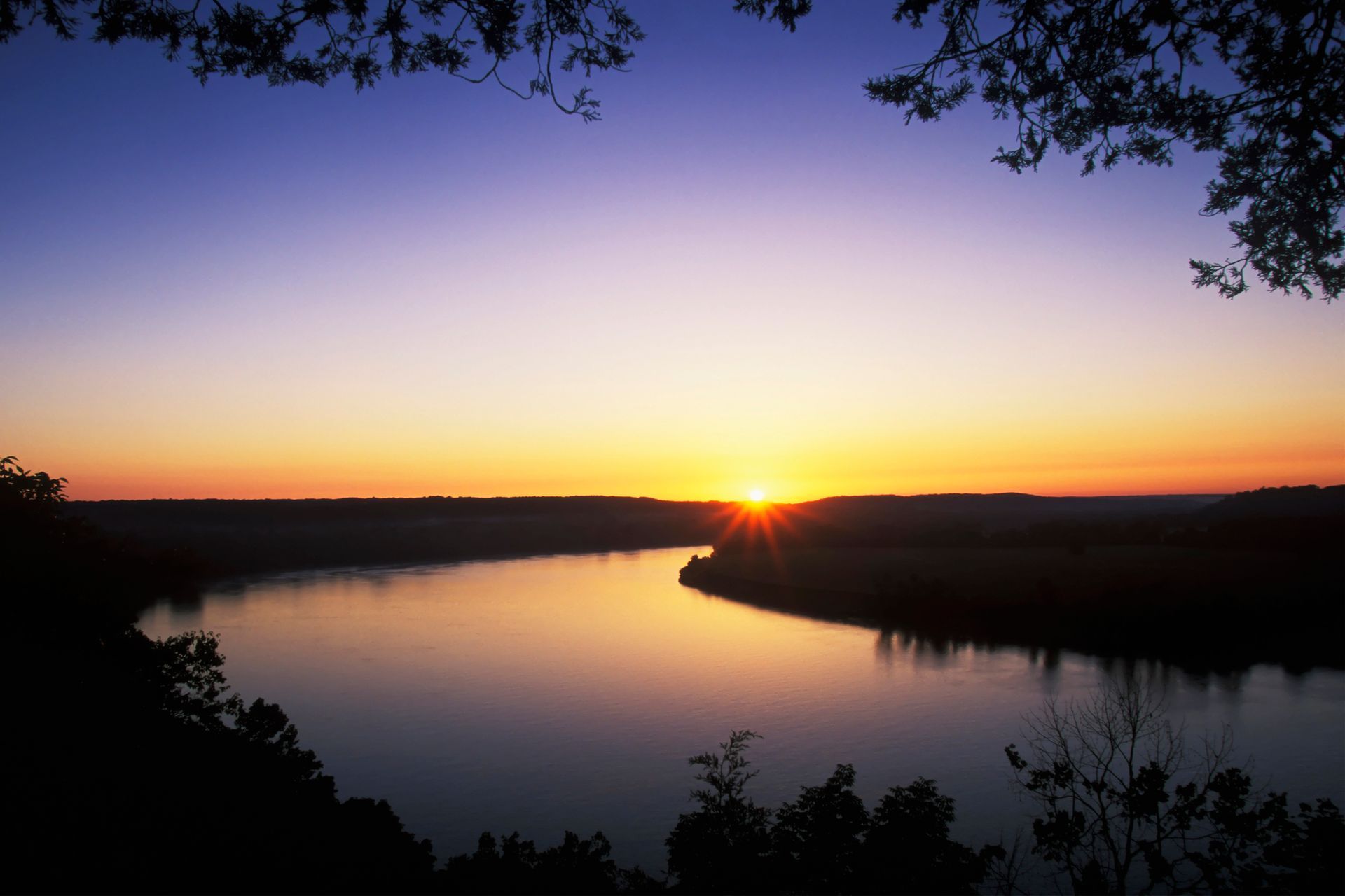The sun is setting over a lake with trees in the foreground.