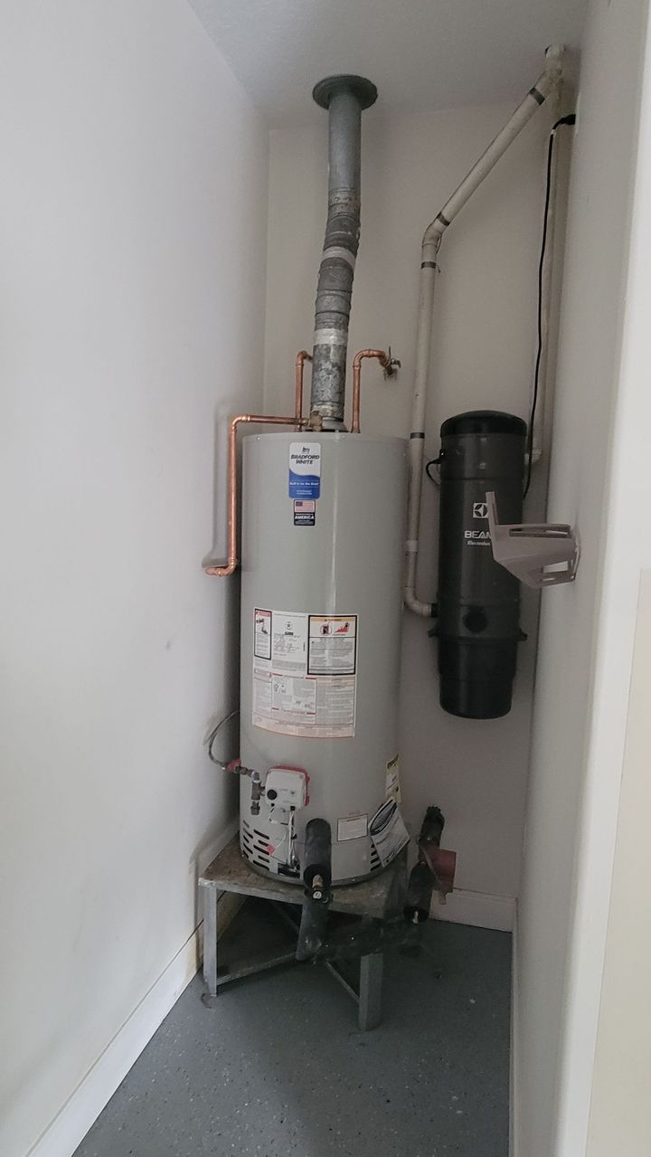 White water heater and black filter tank in a utility closet with exposed pipes on a gray floor