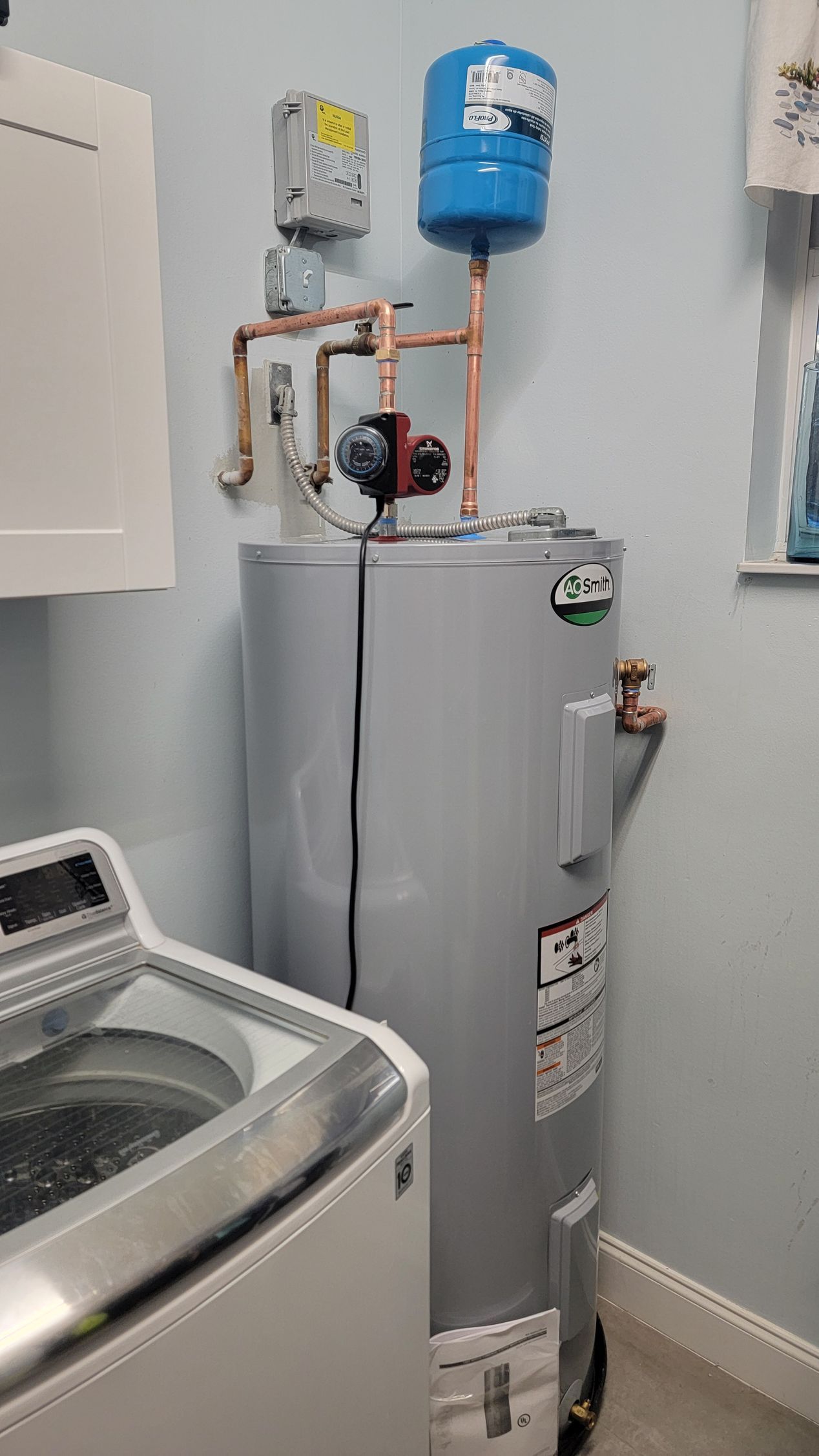 Water heater in a laundry room beside a washing machine, with pipes and a blue expansion tank above.