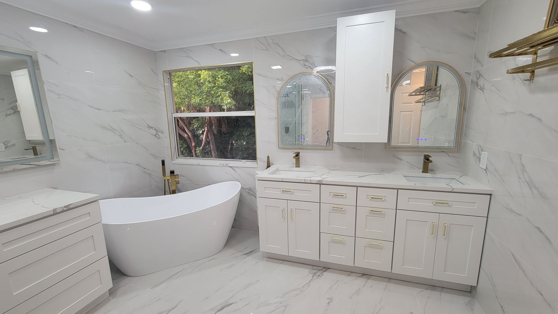 Bright white bathroom with freestanding tub, double vanity, mirrors, and large window overlooking greenery