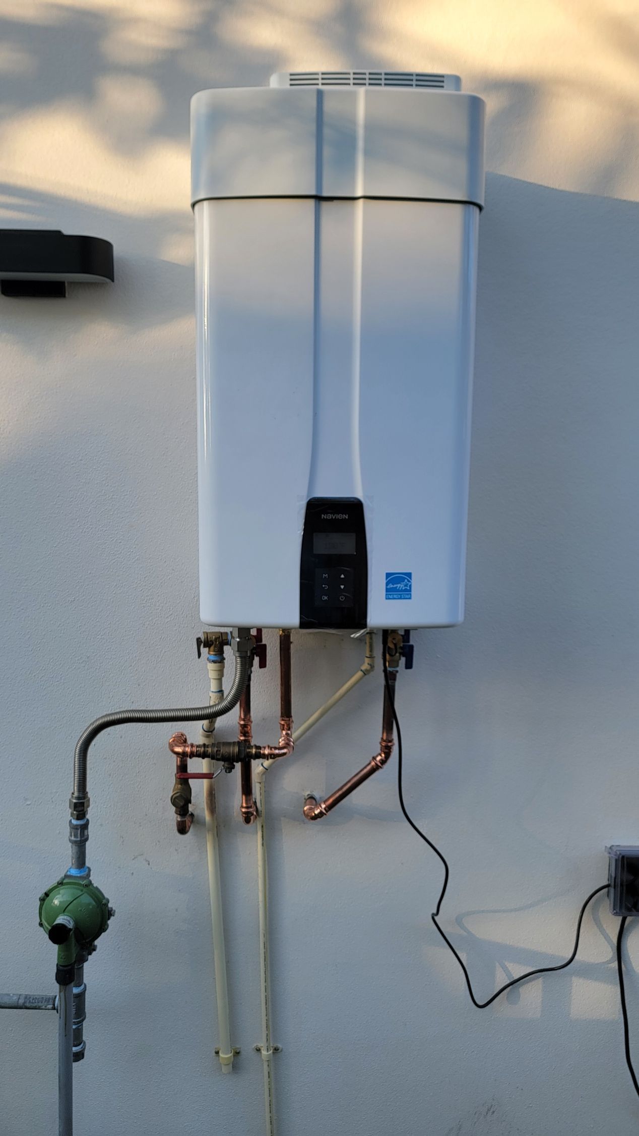Wall-mounted white water heater with pipes and valves against an exterior wall.