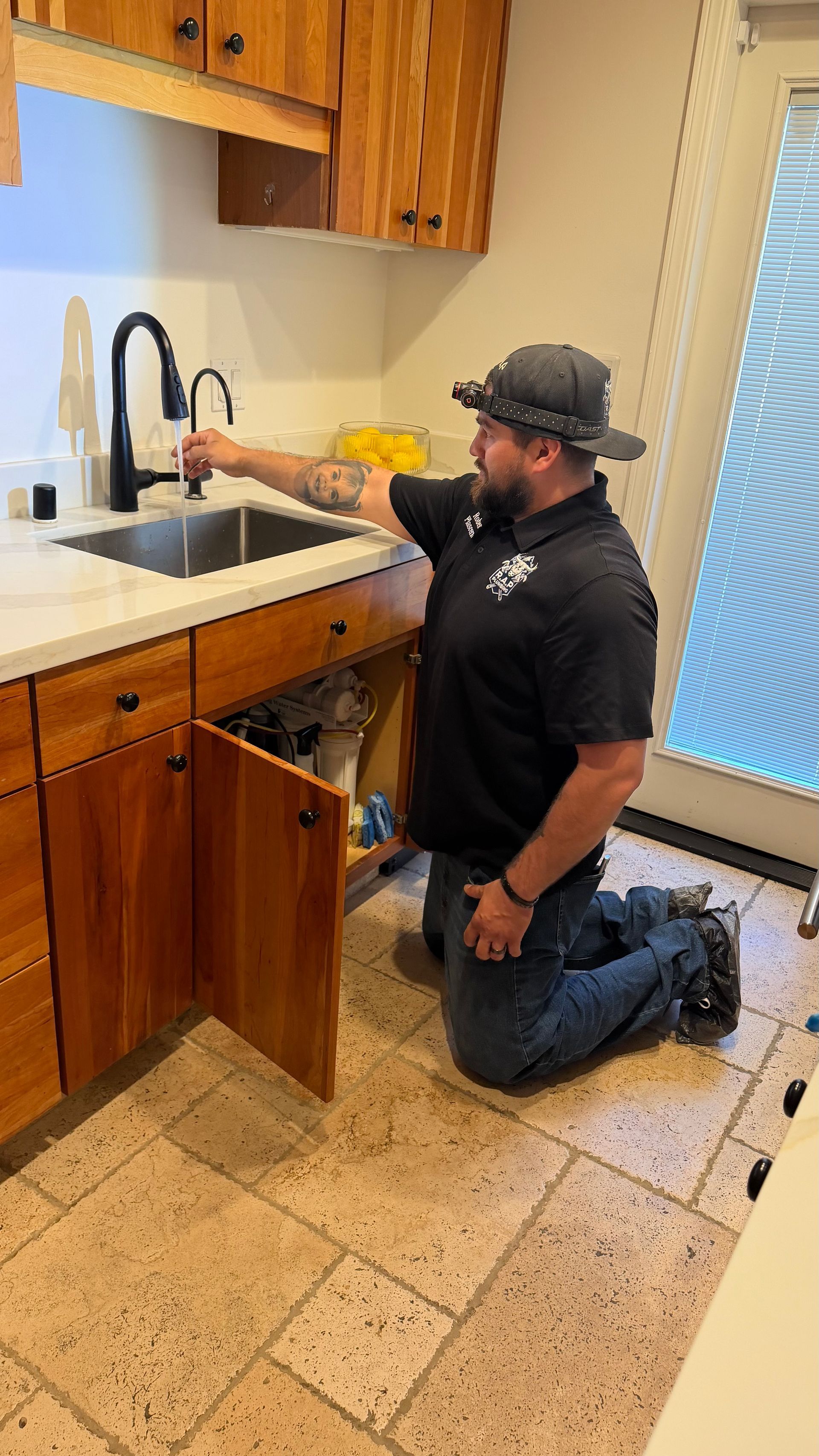 Man kneeling at kitchen sink, reaching toward faucet in a wood-cabinet kitchen.