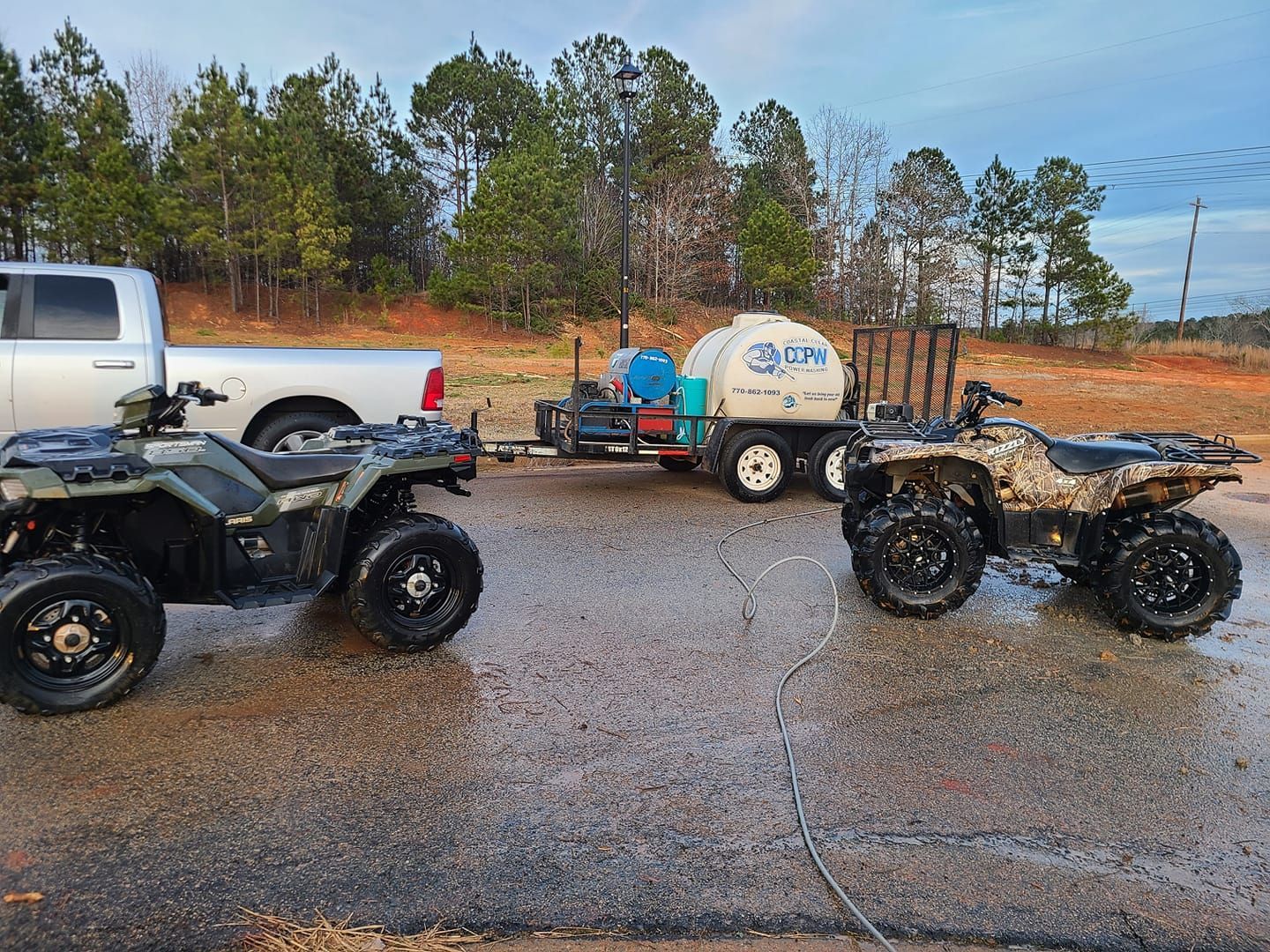 Two ATVs, one olive and one camouflage, parked on a gravel lot in front of a pickup truck pulling a trailer with a tank.