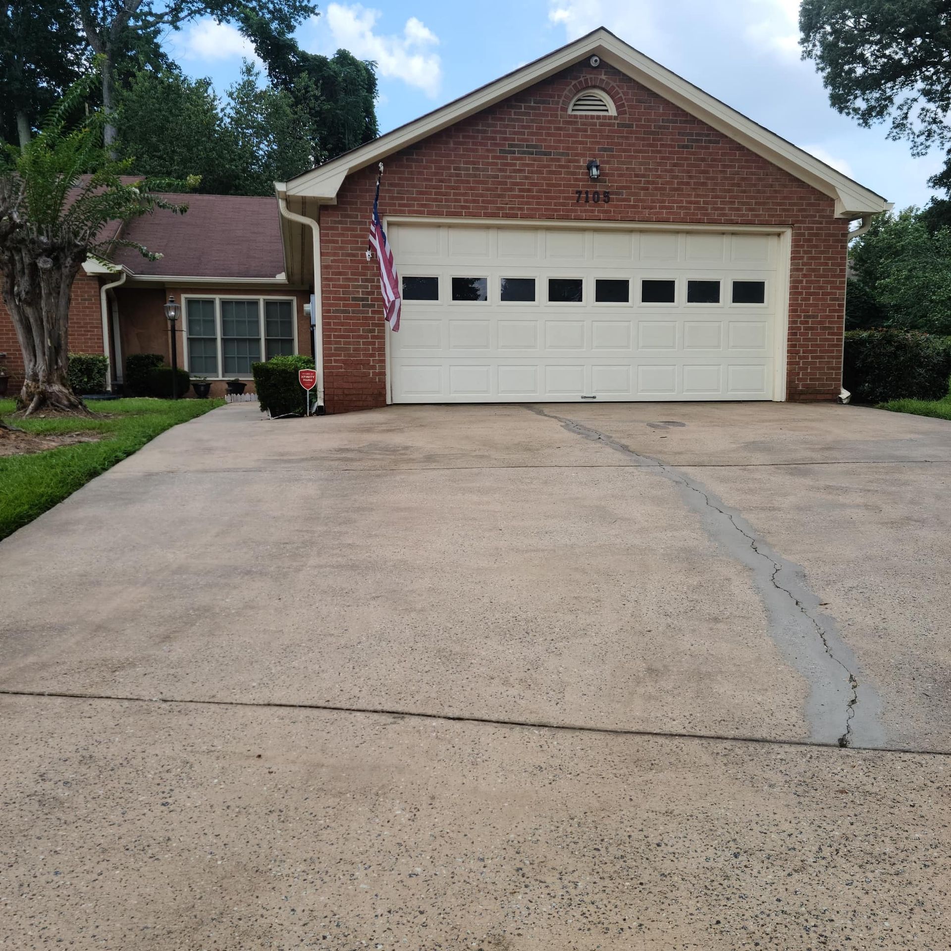 A brick house with a white garage door, an American flag, and a long, weathered concrete driveway.