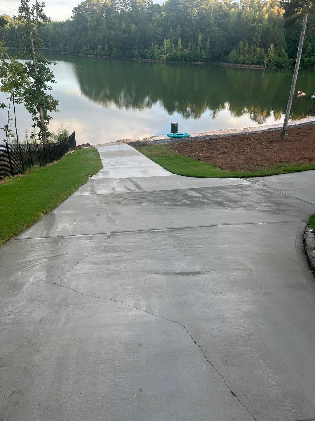 A concrete boat ramp slopes down toward a calm lake surrounded by a dense forest under a bright sky.