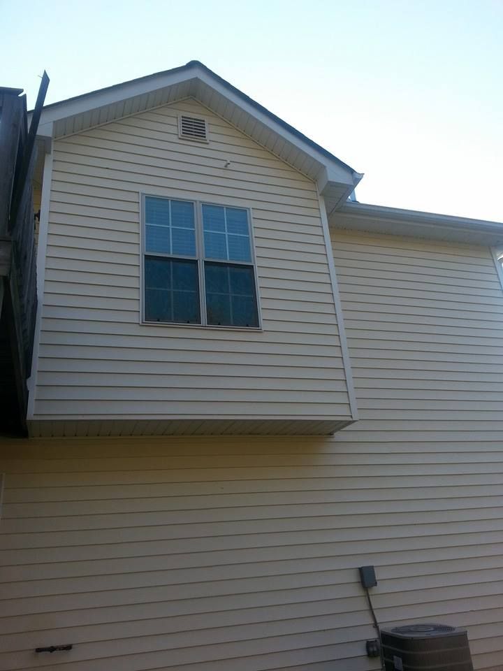 Exterior side view of a house with beige vinyl siding, a protruding upper section, and an outdoor HVAC unit.